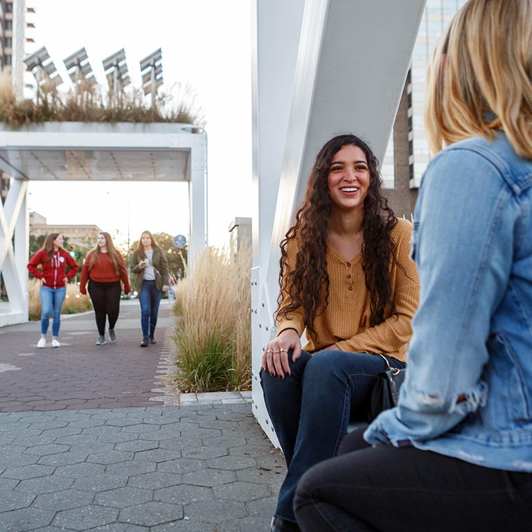 Two students sit on a metal sculpture while three girls walk in the background.